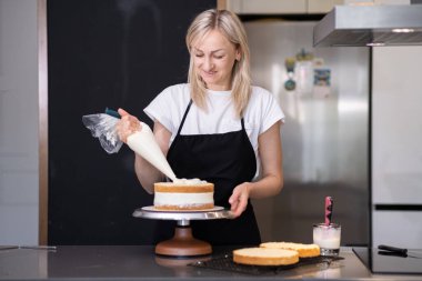 Siyah önlüklü mutlu sarışın kadın yemek blogcusu pasta katmanlarına krem sürerek müşteriler için el yapımı glütensiz şekerleme yapıyor.