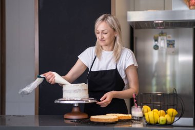 Tatmin olmuş sarışın kadın pastaya organik malzemelerden yapılmış lezzetli bir krema koymak için pasta külahı kullanıyor siyah önlüklü ev hanımı evde el yapımı şekerlemeler yapıyor.