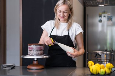Pastaya krema sürmek için siyah önlüklü kadın pastacı. Gülümseyen ve mutfakta ev yapımı şekerleme yaparken eğlenen sarışın kadın.