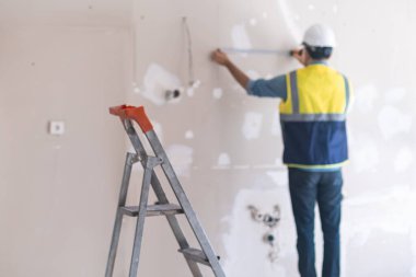 Kurulumdan önce prizler arasındaki mesafeyi belirleyen koruyucu miğferli, pejmürde duvarlı adamı ölçen profesyonel üniformalı bir işçi.