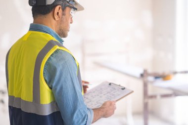 Koruyucu miğfer mühendisi elinde panoyla elinde yenileme projesi olan profesyonel üniformalı bir adam bitmemiş bir odada çizim standı çiziyor.