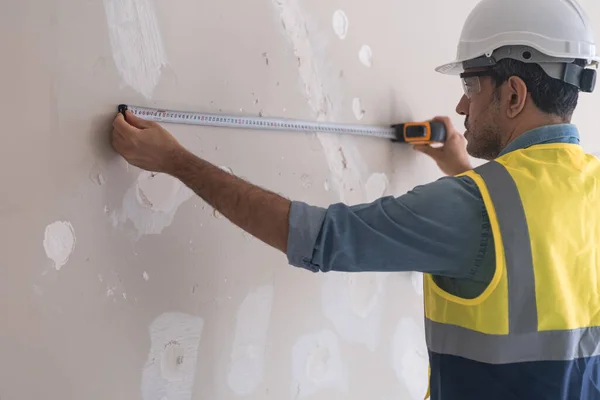 头戴硬礼帽 身穿背心的工人可以测量未来墙壁插座之间的距离 而穿着制服的测量员则负责在公寓客厅里进行翻新 — 图库照片