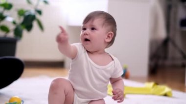 Beyaz elbiseli tombul bir çocuk bembeyaz bir halının üzerine oturmuş bebek odasında çıplak ayakla el hareketleriyle işaret ediyor. Mutlu bir çocukluk ve gelişim dönemi geçiriyor.