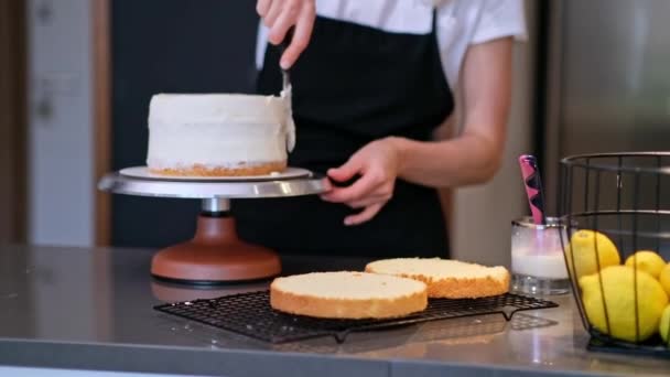フードブロガーの菓子職人は キッチンテーブルに立って黒エプロンで自宅の女性でイベントのためのおいしい乳糖フリーケーキを準備手作りの菓子に白いクリームを笑みを浮かべて実証 — ストック動画