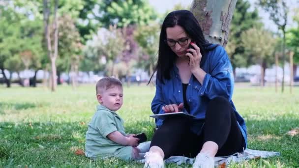在公共花园 一个女人和一个专心致志的儿子坐在树荫下 一边和同事通电话 一边拿着石碑 一边摆出一副镇定的架势 — 图库视频影像