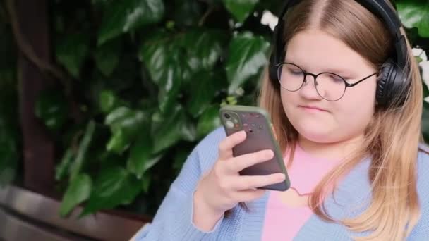 フォーカス十代の女の子でガラスチェックコメントからフォロワーソーシャルネットワーク太りすぎティーンエイジャーでヘッドフォン使用して携帯電話上のベンチに公園に座って — ストック動画