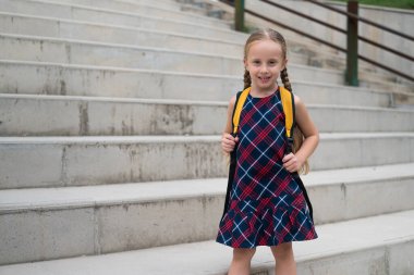 Küçük liseli kız uzun örgülü kameraman kıza gülümsüyor. Elinde okul çantasıyla poz veriyor. Elbiseli hoş bir çocuk parktaki yüksek beton merdivenlerin yanındaki okula gidiyor.