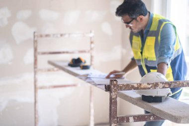 Gözlük konusunda uzman. İnşaat belgelerini inceliyor. Yelekli güvenlik ekipmanının yanında duruyor.