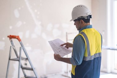 Beyaz kasketli profesyonel mühendis dairedeki tamamlanmamış duvar yenileme sürecinin yanındaki merdivenin önünde el ele duran projeye bakıyor.