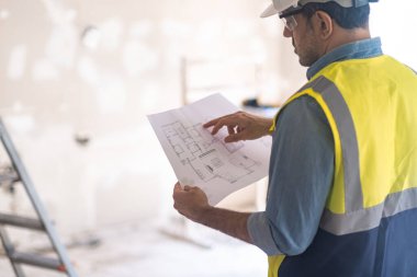Çelik yelek giyen bir adam, yenileme işlemine başlamadan önce dairedeki eski püskü duvarın karşısında duran üniformalı bir uzman tarafından çizim kağıdını kontrol ediyor.