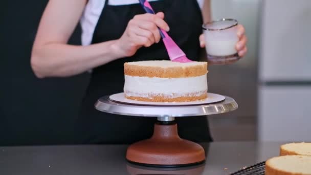 カスタムペストリーを作るテーブルに立って黒エプロンでブロンドの女性の天然成分で作られた有機ケーキの層を塗るためにブラシでガラス瓶から新鮮なクリームを取る濃縮菓子 — ストック動画