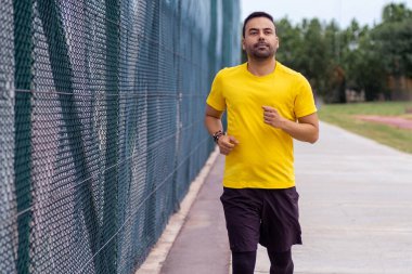 Sakallı, rahat eşofman giyen bir adam, sağlık ve vücut gelişimi için şehir parkındaki spor sahasında yüksek çitleri aşarak yol boyunca koşuyor.