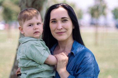Neşeli, koyu saçlı anne, yaz şehrinin bahçesinde küçük bir oğlanın portresini kucaklıyor, esmer kadın ve yakışıklı çocuk güneşli bir günde şehir parkında dinleniyor.