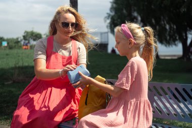 Güneş gözlüklü genç anne, kızına plastik beslenme çantası veriyor. Parktaki bankta oturan kadın, ilkokuldaki derslerden önce okul çocuklarına atıştırmalık veriyor.