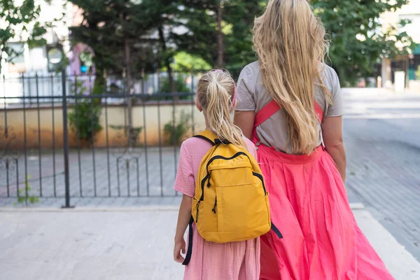 Mother and daughter, backpacks on, walk to school, their figures seen from behind.