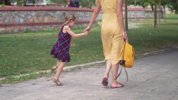 小さな女の子は 黄色い夏のドレスで緑の公園の若い女性を横断して母と歩く道路タイルで遊びます アクティブな娘を市の庭の子供の世話で学校に連れて行きます — ストック動画