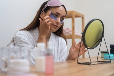 Bornozlu çekici kadın göz altı bantları uyguluyor. Masadaki yuvarlak aynaya bakıyor. Güzel bayan spa gününde yaşlanma karşıtı cilt bakımı yapıyor.