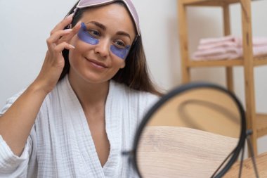 Puffy-Eye Mükemmellik: Aynanın yansımasında, bir kadın güne şişkinlik, kırışıklıklar ve koyu halkalarla savaşmak için göz altı maskeleri takarak başlar. 