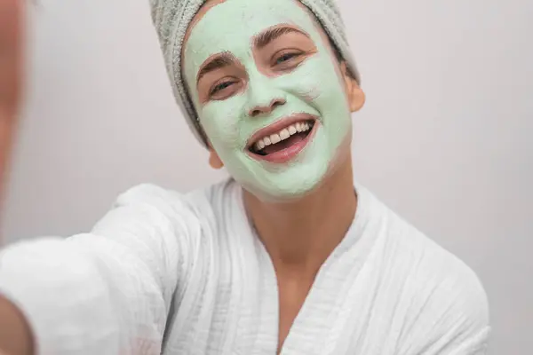 Joyful woman with clay mask on face wearing terry bathrobe making selfie on white background closeup happy lady recording beauty blog about skincare at home