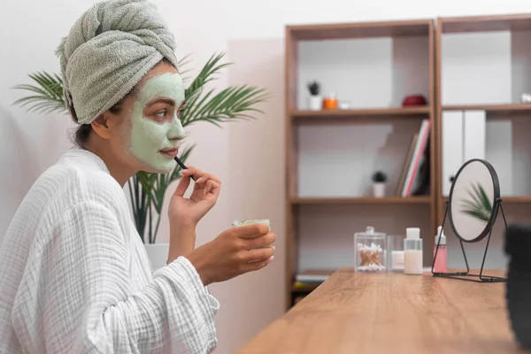 young woman applying green clay mask on face looking in table mirror in bathrobe organic cosmetics at home side view