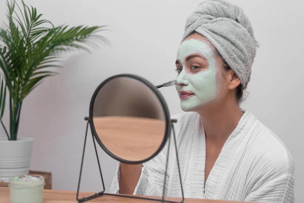 Happy woman spreading green clay mask with brush on facial skin looking in table mirror lady in gown enjoying face cleansing procedure on pampering day