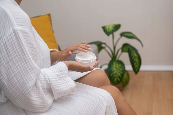 Bathrobe beauty: At home, a woman nourishes her skin with moisturizing lotion from a jar, elevating her body care routine.