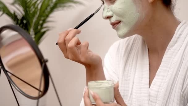 自家製粘土マスクを顔面に塗布する女性は 自宅で顔の皮膚を浄化するためにクラフトオーガニック化粧品を使用してバスローブのテーブルミラーレディに座ってブラシをかけます — ストック動画