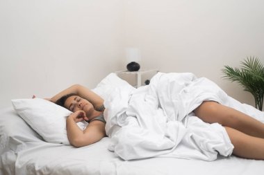 Gecelikli kadın yatakta rahat bir şekilde uyuyor. Yatak odasında pastel renkler ve apartmandaki saksı bitkisi olan kadın uykusundan enerji alıyor.