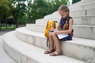 Yanında sırt çantası olan sevimli bir liseli kız okul bahçesindeki taş basamaklarda otururken dikkatle defterine yazıyor.. 