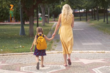 Genç bir kız, her ikisi de sırt çantasıyla okula giderken annesinin yanında mutlu bir şekilde zıplıyor.. 