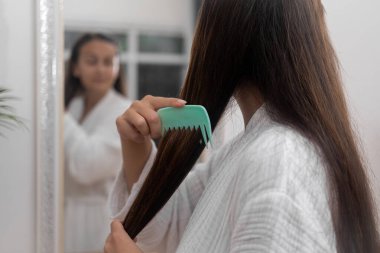 Uzun düz kestane rengi saçları olan bir kadın banyoda aynaya bakıyor bornozlu kadın evde saç bakımının keyfini çıkarıyor.