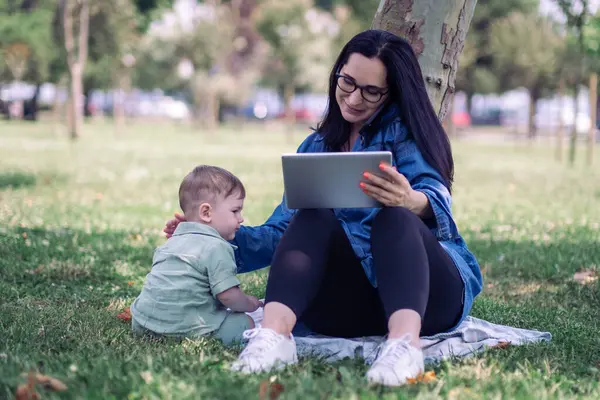 Businesswoman consulting client by phone and working on tablet while playing with baby boy under tree on green park lawn woman freelancer with son outdoors