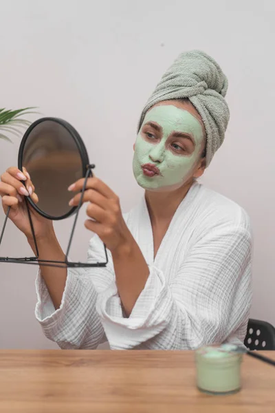 Woman with green clay mask on face and towel wrap on head sending kiss to mirror reflection at table lady in bathrobe peeling facial skin on home pampering day