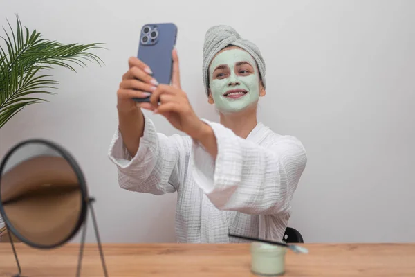 Blogger Beauty: In her bathrobe, she dons a natural clay mask, capturing skincare content for social media with a selfie in her home studio.