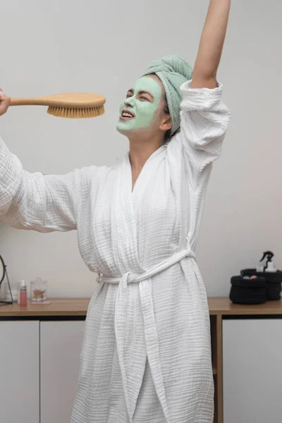 Funny young woman with cleansing clay mask on face singing in wooden hairbrush at home joyful lady in bathrobe enjoying skincare treatment on pampering day