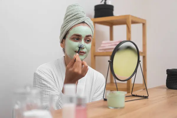 Positive woman applying green clay mask on nose with brush looking in table mirror young lady in terry bathrobe doing face cleansing procedure at home