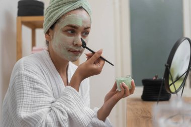 Ev Spa Serenity: Bornozuyla, organik yüz tedavisinden zevk alıyor, aynaya bakarken kilden bir maske takıyor.. 