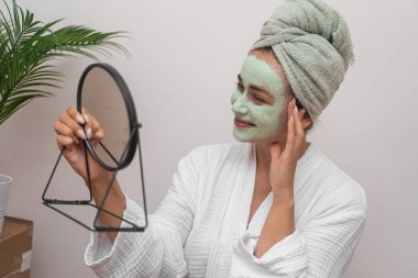 Maskeli Güzel: Neşeli bir kadın, cüppeli, aynaya bakarken gülümseyen, yenilenen bir cilt için ev yapımı yeşil kilden bir maske kullanan. 