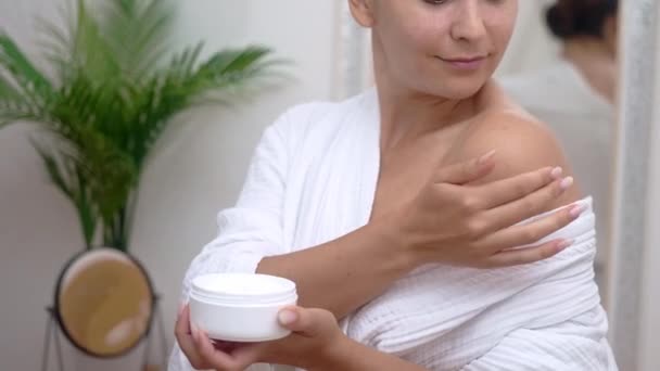 在浴室里 穿着浴衣的漂亮女士在裸露的肩膀上涂润肤霜 在家里用化妆品进行有效的身体护理 — 图库视频影像