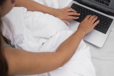 Kadın sosyal ağlarda internet üzerinden iletişim kuruyor evde yatakta kadın elleri beyaz battaniyeye uzanıyor ve akşamları dizüstü bilgisayarın klavyesine mesaj yazıyor.