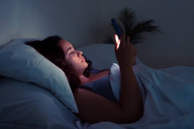 Sosyal ağlarda arkadaşlarıyla sohbet eden genç ve çekici bir bayan karanlık bir yatak odasında uzanmış cep telefonu ekranına bakıyor ve uyumadan önce dinleniyor.