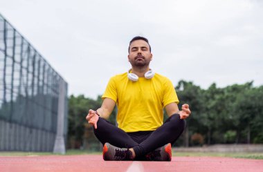 Canlı spor kıyafetleri içindeki sakin sporcu bir stadyumdaki koşu pistinde Lotus pozunda meditasyon yapıyor..