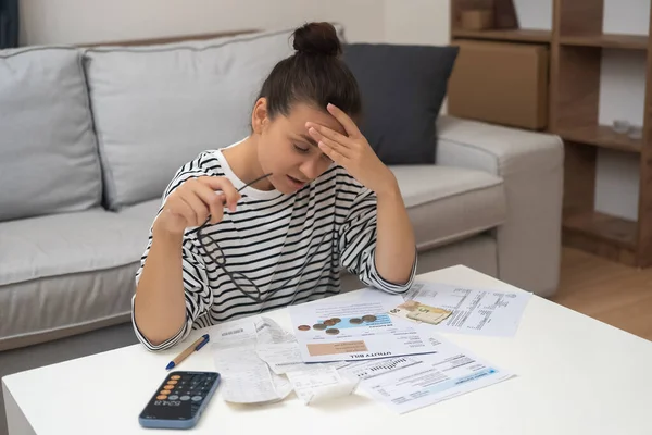 Woman carefully examines papers with prices of utilities for month lady accountant counts total amount holding head with hand sitting at coffee table in light flat