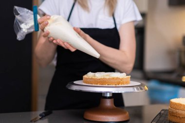 Mutfak masasındaki siyah önlüklü kadının üzerine taze krema koymak için pasta konisi kullanan profesyonel bir pastacı. Doğal malzemelerden lezzetli pastalar yapıyor.