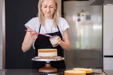 Konsantre şekerlemeci cam kavanozdan taze krema alıyor ve doğal malzemelerden yapılmış organik pasta katmanları sürüyor. Siyah önlüklü sarışın kadın masada özel pasta yapıyor.