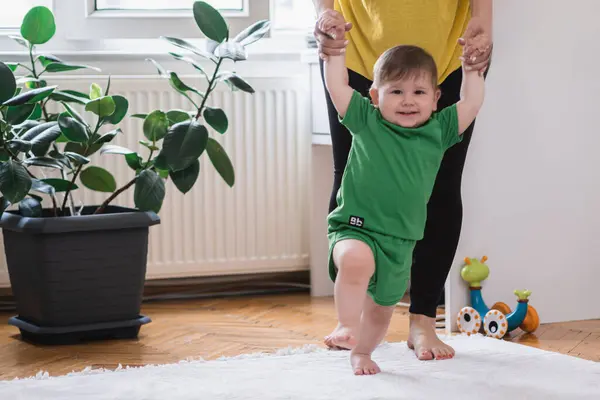 Happy little boy in green outfit holding mother hands learning to make first steps woman teaching toddler son to walk on soft rug put near houseplant in living room at home