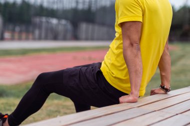 Rahat giysiler içinde güçlü bir adam geriye doğru şınav çekiyor. Şehir parkındaki sporcuların rutin antrenmanlarında ahşap bankta yaslanıyor. Boş spor sahasının kapanış sahnesinde.