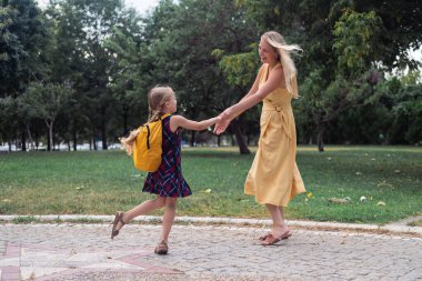 Ateşli genç bir kız öğrenci, sarı sırt çantası zıplayan, okula giderken annesinin yanında mutlu bir şekilde zıplıyor.. 