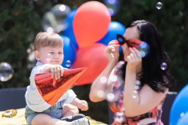 Vibrant one-year-old party - baby wears a festive Happy Birthday hat while enjoying an outdoor celebration featuring red and blue balloons, playful soap bubbles