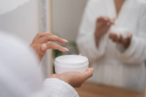 Woman hands holding lotion jar at dressing table in bathroom lady in gown taking anti aging skincare product from plastic bottle for applying in bathroom closeup